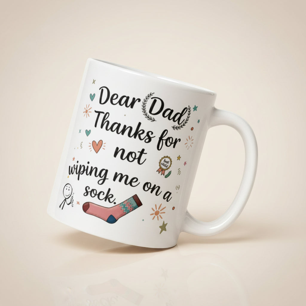 A tilted white ceramic mug on a light beige surface. The design features playful, hand drawn style text that reads DEAR DAD THANKS FOR NOT WIPING ME ON A SOCK. The typography is surrounded by whimsical icons including a pink patterned sock, a smiling stick figure, a gold best child medal, and small sun and heart illustrations.