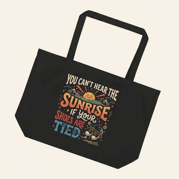 A large black eco tote bag featuring a retro 1970s design of a smiling sun and mountains with the funny, nonsensical quote You Can Not Hear The Sunrise If Your Shoes Are Tied.