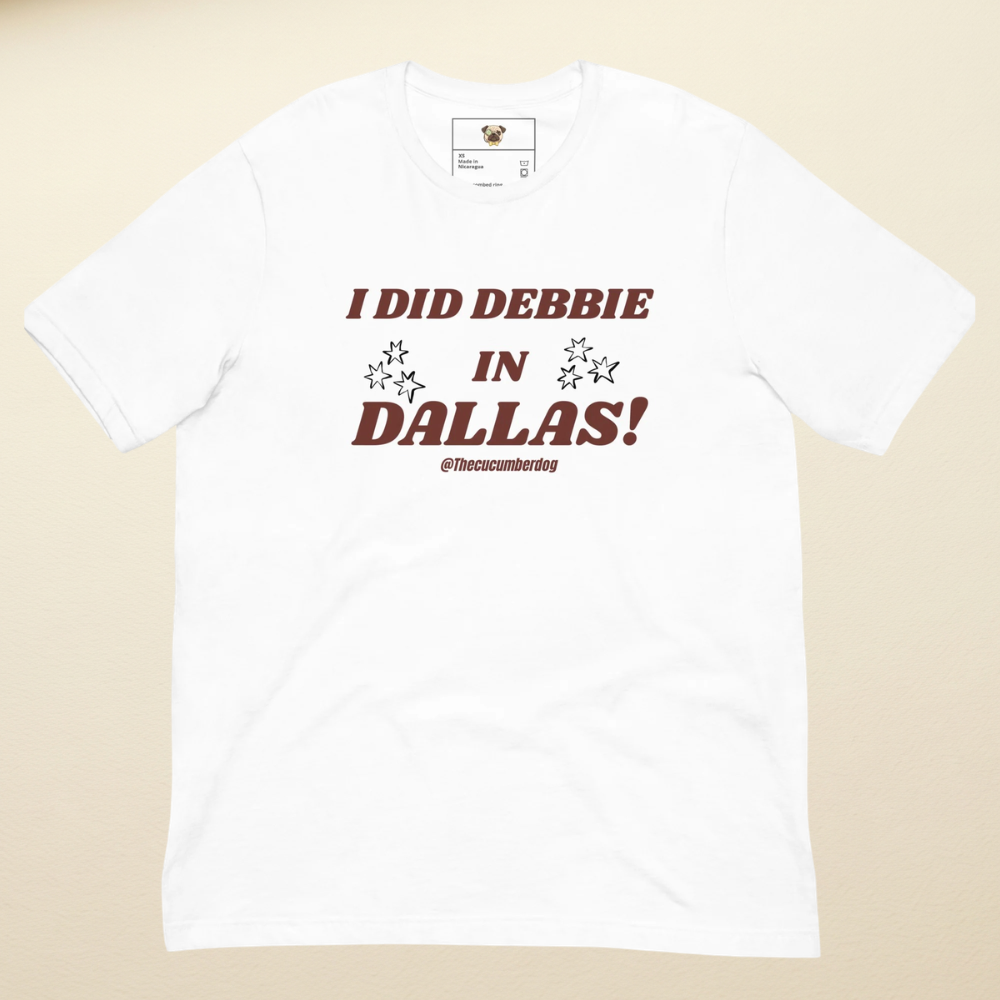 A white short-sleeve crewneck t-shirt with a graphic printed on the chest reading 'I DID DEBBIE IN DALLAS!l  and the handle '@thecucumberdog' below. The shirt is laid flat against a light beige background.