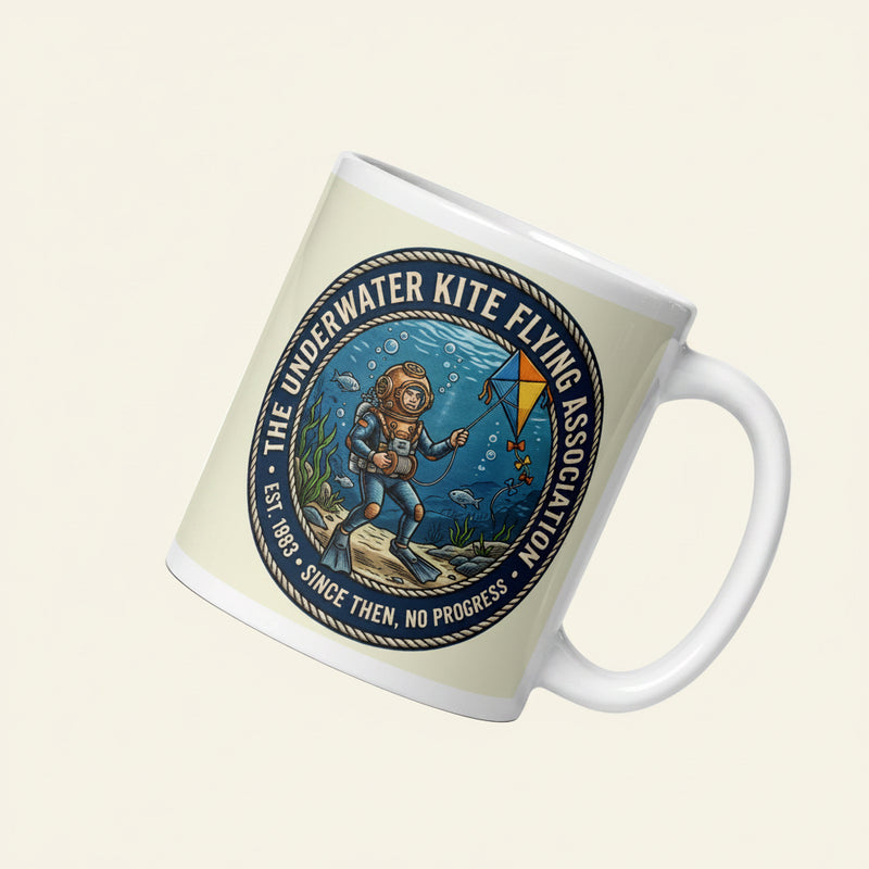 A white ceramic mug featuring a retro illustration of a vintage deep sea diver underwater attempting to fly a blue and yellow kite among fish and seaweed. The circular nautical rope border reads The Underwater Kite Flying Association, Est. 1983, Since then, no progress.