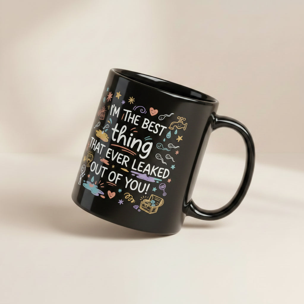 A tilted black glossy ceramic mug on a light beige background. The design features colourful hand drawn typography and doodles. The text reads I'M THE BEST thing THAT EVER LEAKED OUT OF YOU! in white and teal script. The surrounding icons include white sperm cells, a dripping golden tap, hearts, stars, a treasure chest, and a baby rattle in pastel colours.