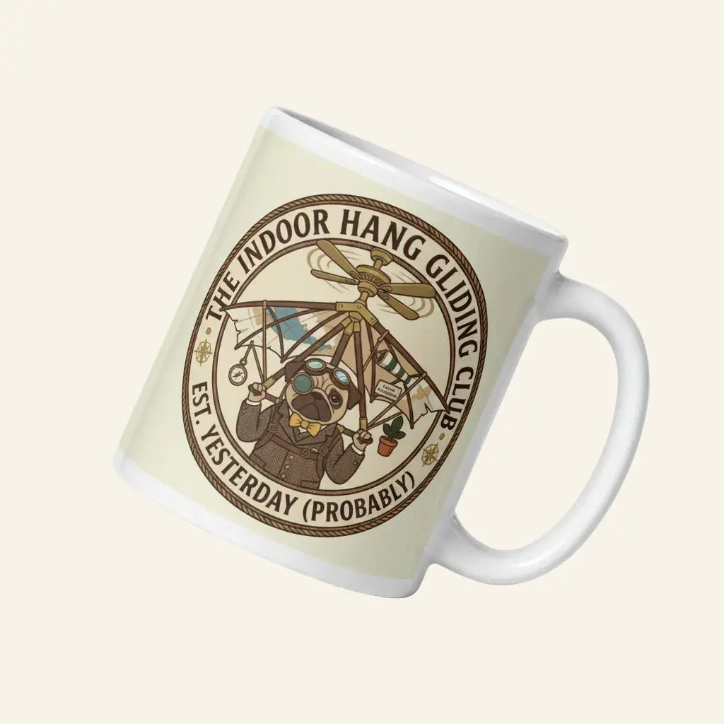 A circular vintage emblem on a transparent background. It reads THE INDOOR HANG GLIDING CLUB EST. YESTERDAY (PROBABLY). Inside, Sir Cucumber Dog wears a tweed suit and aviator goggles, strapped to a hang glider that is attached to a ceiling fan. 11 oz