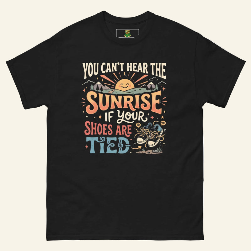 A black graphic t-shirt featuring a smiling sun rising over mountains and the sarcastic quote You Can’t Hear the Sunrise if Your Shoes are Tied in vintage typography with a pair of untied blue sneakers below. Cucumber Dog