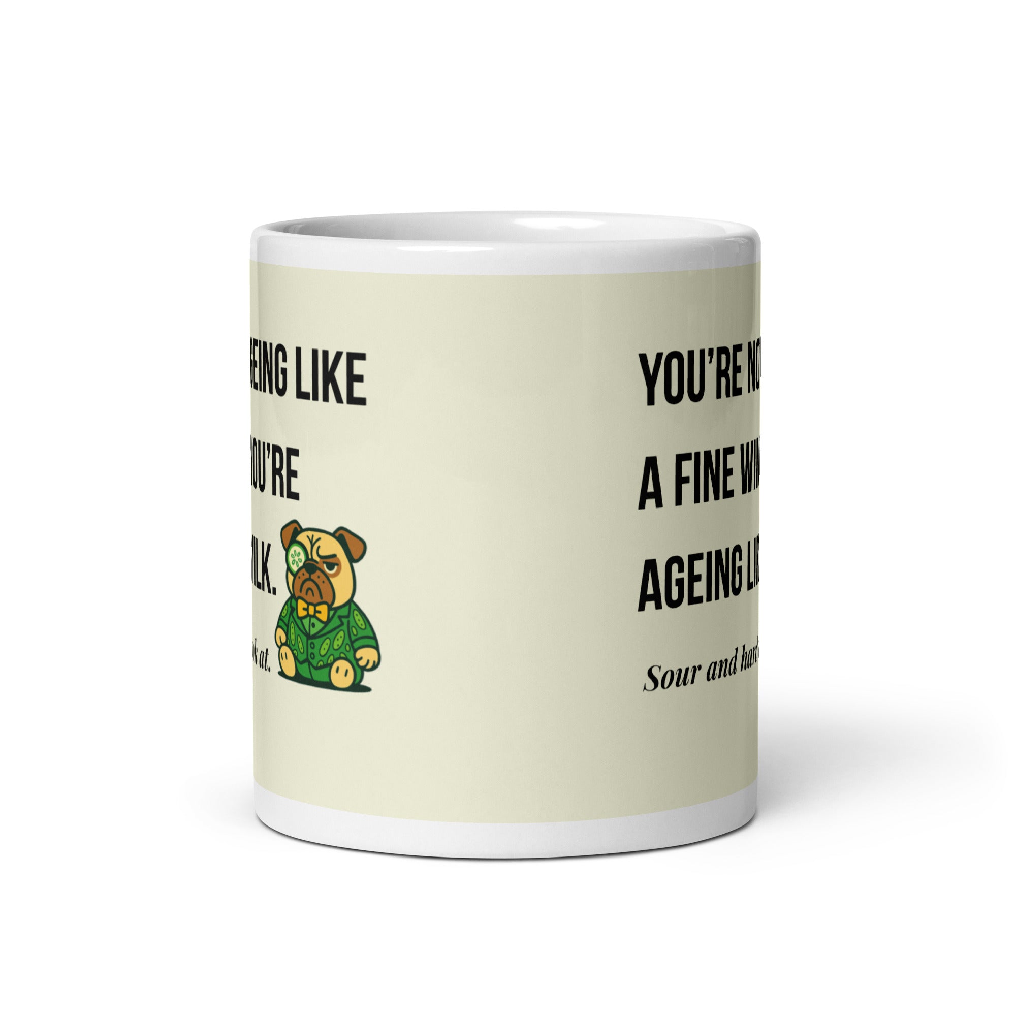 White glossy mug featuring Sir Cucumber Dog the pug. The text "YOU'RE NOT AGEING LIKE A FINE WINE. YOU'RE AGEING LIKE MILK. SOUR AND HARD TO LOOK AT." is rendered in Cucumber green over a vibrant cream background banner.