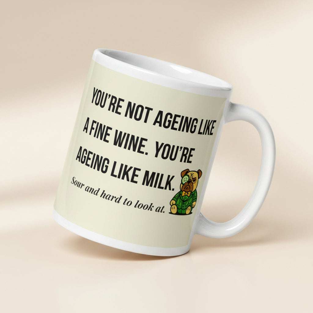 White glossy mug featuring Sir Cucumber Dog the pug. The text "YOU'RE NOT AGEING LIKE A FINE WINE. YOU'RE AGEING LIKE MILK. SOUR AND HARD TO LOOK AT." is rendered in Cucumber green over a vibrant cream background banner.