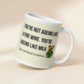 White glossy mug featuring Sir Cucumber Dog the pug. The text "YOU'RE NOT AGEING LIKE A FINE WINE. YOU'RE AGEING LIKE MILK. SOUR AND HARD TO LOOK AT." is rendered in Cucumber green over a vibrant cream background banner.