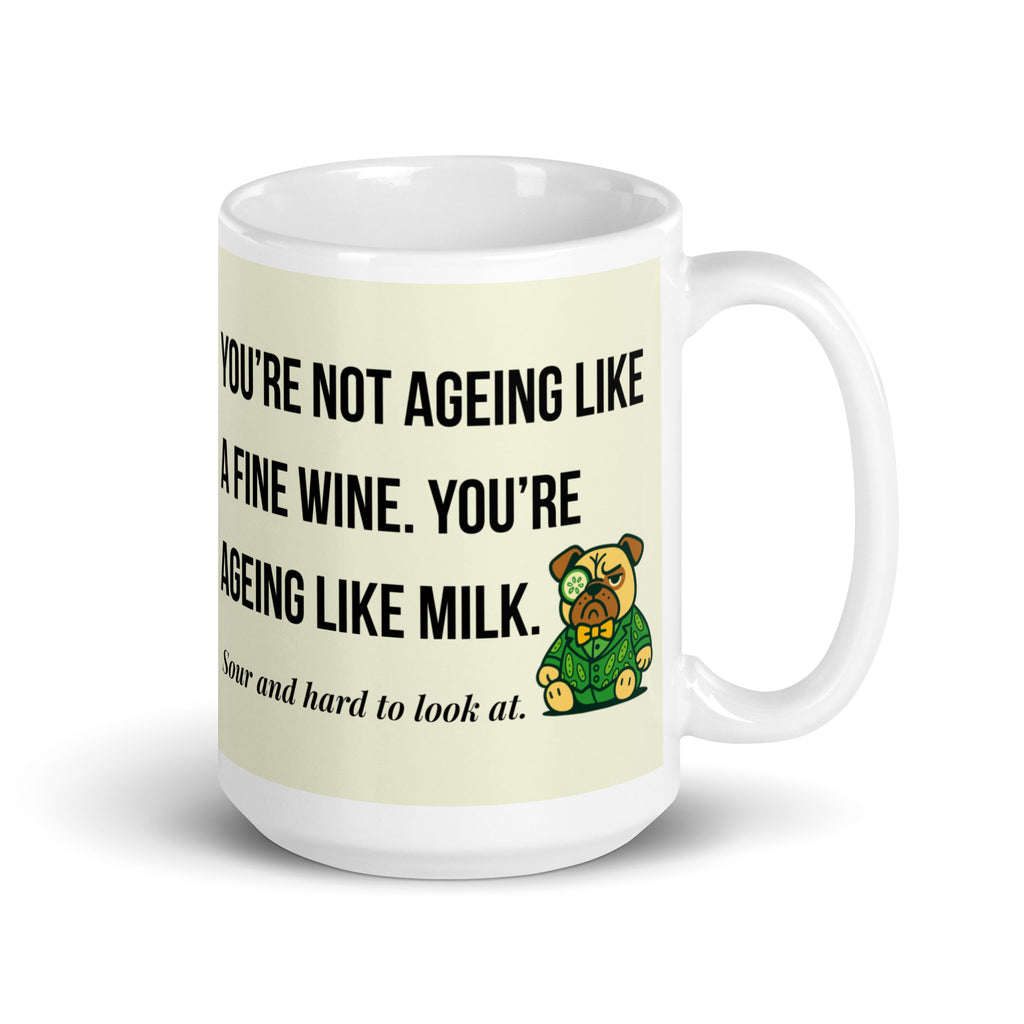 White glossy mug featuring Sir Cucumber Dog the pug. The text "YOU'RE NOT AGEING LIKE A FINE WINE. YOU'RE AGEING LIKE MILK. SOUR AND HARD TO LOOK AT." is rendered in Cucumber green over a vibrant cream background banner. 15oz